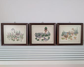 May include: Three rectangular wall hangings featuring cross-stitch embroidery of birds and waterfowl. Each piece is enclosed in a dark brown wooden frame with a metal hanging loop. The needlework depicts geese, ducks, and pigeons in natural settings.