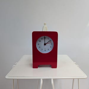 May include: A red rectangular clock with a white clock face and black numerals. The clock has a white top and is sitting on a white metal side table. The time on the clock is approximately 1:07.