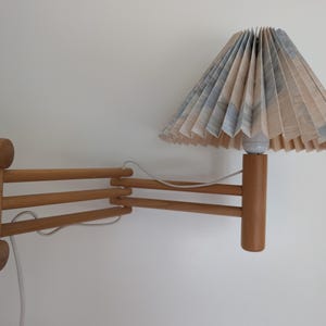 May include: A wall-mounted lamp featuring an extendable wooden scissor arm mechanism and a pleated conical lampshade. The shade displays a subtle blue and beige pattern. A white electrical cord runs along the wooden arm to the light socket.
