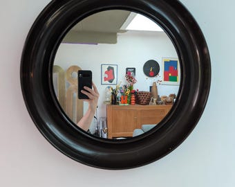 Pode incluir: Um espelho de parede circular com uma moldura preta larga, lisa e com acabamento brilhante. A superfície refletora mostra o interior de uma sala, incluindo um aparador de madeira, vários objetos decorativos e uma mão segurando um smartphone preto.