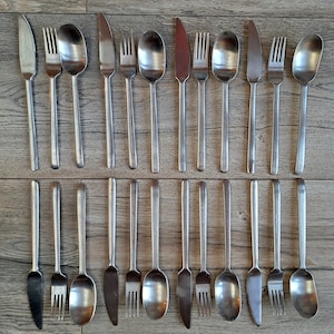 May include: An assortment of stainless steel cutlery, comprising knives, forks, and spoons, displayed on a wooden surface. The cutlery features a straightforward, timeless design with elongated, slim handles. The arrangement implies a formal dining occasion.