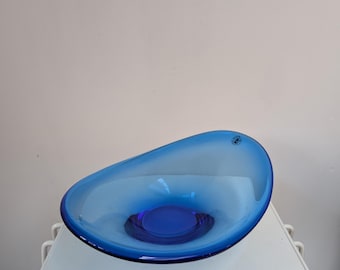 Pode incluir: Uma taça de vidro azul translúcido com uma forma orgânica assimétrica. A taça apresenta um centro azul cobalto profundo que transita para um tom azul mais claro e vibrante em direção à borda. Um pequeno autocolante circular é visível.