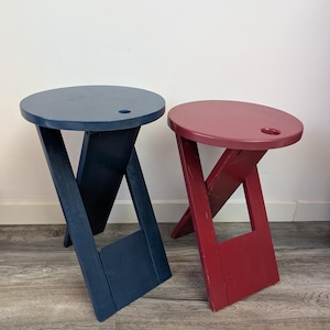 Op de afbeelding: Twee houten klapstoelen, één marineblauw, de andere rood. Elk heeft een ronde zitting met een klein gat en schuine poten. De krukken zijn ontworpen om op te vouwen voor gemakkelijke opslag. Ruimtebesparend en draagbaar ontwerp.