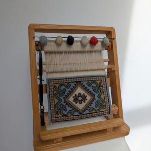 Op de afbeelding: Een houten weefgetouw met een klein tapijt erop geweven. Het weefgetouw heeft een standaard en een set kleurrijke garenbollen die aan de bovenkant hangen.