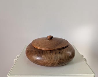 May include: A round, dark brown wooden lidded bowl with a smooth, polished finish and a small, central knob handle on the lid. The wood grain is visible across the surface of the bowl and the lid, showcasing natural patterns and textures.