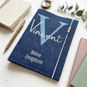 Könnte beinhalten: Eine dunkelblaue Filzmappe mit einem großen hellblauen Buchstaben V, dem Namen Vincent in weißer Schreibschrift und dem Text Meine Zeugnisse in weißen Druckbuchstaben. Ein weißes Gummiband mit drei kleinen Holzperlen verschließt die Mappe.