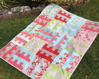 May include: A patchwork quilt with a geometric pattern in shades of pink, red, green, and blue. The quilt is made of various floral fabrics and is laid out on a grassy surface.
