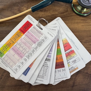 Könnte beinhalten: Ein Satz weißer medizinischer Referenzkarten, die durch einen Metallring zusammengehalten werden und verschiedene medizinische Diagramme und Grafiken anzeigen. Ein Stethoskop liegt in der Nähe auf einer Holzoberfläche. Die Karten enthalten Informationen zu V-Tach, PEA usw.