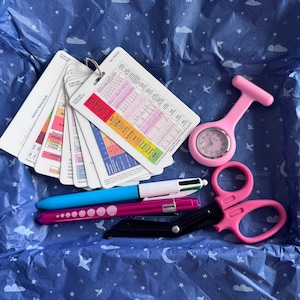 May include: A set of medical reference cards on a metal ring, a pink silicone fob watch, a multi-colour ink pen, a magenta pen with white dots, and black trauma shears with pink handles. The cards feature various charts, text, and colour-coded sections.