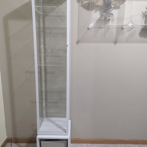 Puede incluir: Una vitrina de cristal blanca con un caj&oacute;n en la parte inferior. La vitrina tiene cinco estantes y est&aacute; hecha de cristal transparente y metal blanco.