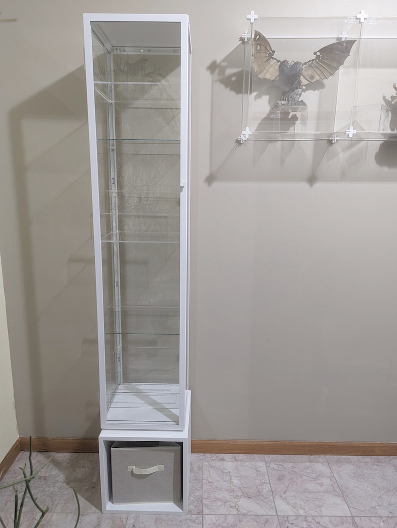 Puede incluir: Una vitrina de cristal blanca con un caj&oacute;n en la parte inferior. La vitrina tiene cinco estantes y est&aacute; hecha de cristal transparente y metal blanco.