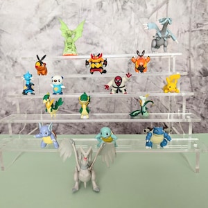 Op de afbeelding: Een verzameling kleurrijke Pokémon-beeldjes tentoongesteld op een heldere, gelaagde standaard. De beeldjes tonen verschillende personages in levendige kleuren, waaronder blauw, groen, geel en oranje. De presentatie is tegen een neutrale achtergrond.