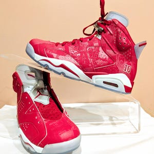 May include: A pair of red and white Air Jordan 6 Retro sneakers with a white sole and a red upper. The sneakers have a white and red pattern on the upper and the number "10" is printed on the side of the right shoe.