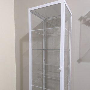 Puede incluir: Vitrina de cristal blanca con cinco estantes. La vitrina tiene una puerta de cristal transparente y un marco blanco.
