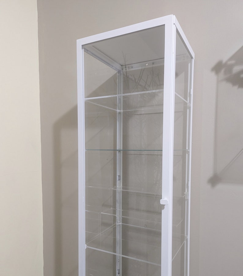 Puede incluir: Vitrina de cristal blanca con cinco estantes. La vitrina tiene una puerta de cristal transparente y un marco blanco.