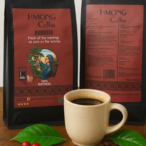 以下が含まれることがあります： 「HMONG Coffee Robusta」と書かれた赤いラベルが付いた2つの黒いコーヒーバッグ。コーヒー豆、赤いベリー、緑の葉が入ったベージュのコーヒーマグが前にあります。バッグにはイラストとテキストがあります。