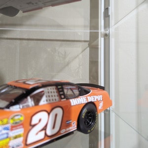 Puede incluir: Coche de carreras naranja y blanco con el n&uacute;mero 20 en el lateral y "Home Depot" escrito en la parte trasera. El coche se exhibe en una vitrina.
