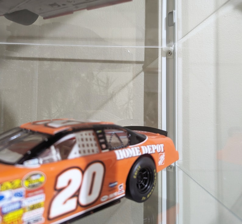 Puede incluir: Coche de carreras naranja y blanco con el n&uacute;mero 20 en el lateral y "Home Depot" escrito en la parte trasera. El coche se exhibe en una vitrina.