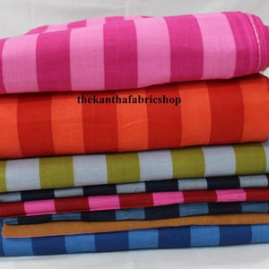 May include: A stack of folded fabric bolts featuring various striped patterns in vibrant colors including hot pink, orange, olive green, navy blue, and light blue. The text thekanthafabricshop is printed in black across the orange fabric layer.