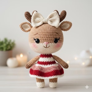 Peut inclure: Poupée cerf au crochet faite à la main avec un nœud crème, des bois bruns et une robe rayée rouge, rose et blanche. La poupée a un corps brun clair, des joues roses et des yeux noirs. La poupée est sur une surface blanche.