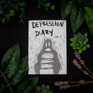 Könnte beinhalten: Ein kleines, weißes Buch mit dem Titel "DEPRESSION DIARY Vol. I" in schwarzem Stift geschrieben. Der Umschlag zeigt eine Schwarzweißillustration einer Figur mit verdecktem Kopf, die mit Seilen gebunden ist.