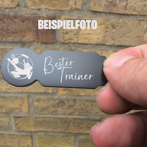 Puede incluir: Un llavero rectangular gris oscuro con un diseño temático de fútbol y las palabras "Bester Trainer" en escritura blanca. El llavero presenta a un jugador de fútbol pateando un balón. La palabra "BEISPIELFOTO" está en el fondo.