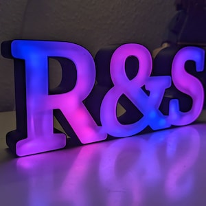 May include: A light-up sign with the letters "R&S" in black with a white interior. The "R" is lit with a pink and blue gradient, and the "S" is lit with a blue and purple gradient.