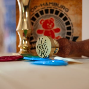 May include: A brass wax seal stamp with a wooden handle, lying on a surface with colorful wax seals. A gold-colored decorative item and a vintage-style sign with a teddy bear graphic are in the background.