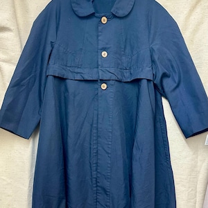 May include: A navy blue coat with a round collar and three wooden buttons. The coat has three-quarter length sleeves and a unique design element across the chest. The coat is hanging on a clear hanger.