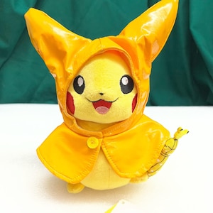 May include: A yellow Pikachu plush toy wearing a bright yellow raincoat with a hood and cape. The plush has black eyes, red cheeks, and a smiling mouth. It is holding a yellow and white umbrella.