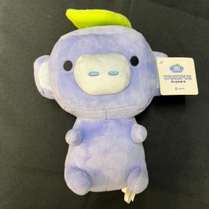 May include: A plush toy in the shape of a cartoon character, primarily lavender with a white snout and ears. It has black eyes and a small green leaf on its head. The tag reads "Wumpus Plushie".