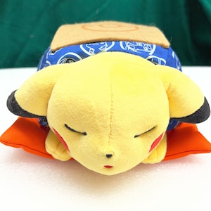 May include: A yellow Pikachu plush toy with black-tipped ears, red cheeks, and closed eyes, resting on an orange pillow. It has a blue patterned covering and a tan square on its back. A cute, soft toy.