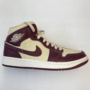 May include: A high-top sneaker in cream and burgundy leather. The shoe features a white midsole and a burgundy outsole. The iconic Nike swoosh is visible on the side, and the Air Jordan logo is on the ankle.