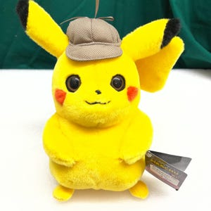 May include: A plush Pikachu toy, primarily yellow with black-tipped ears and red cheek markings, wearing a small brown tweed detective hat. The toy has a tag attached. The background is a mix of white and green.