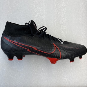 May include: Black football boot with red detailing, featuring a high-top design. The Nike swoosh logo is red on the side. The boot has a textured upper and red studs on the sole.