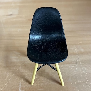 May include: A miniature black chair with a textured seat and back. The chair has four yellow legs and a black metal support structure. The chair is a replica of a classic mid-century modern design.