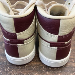 May include: Pair of high-top sneakers in cream and burgundy. The shoes feature a color-blocked design with cream-colored panels and burgundy accents on the heel and side. The soles are white, and the laces are cream-colored.