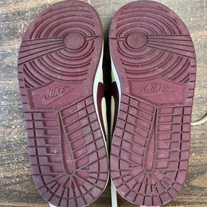 May include: Pair of Nike sneakers, showcasing the soles. The soles are a deep burgundy color with a textured pattern and the Nike logo. The uppers are a cream color with burgundy accents.