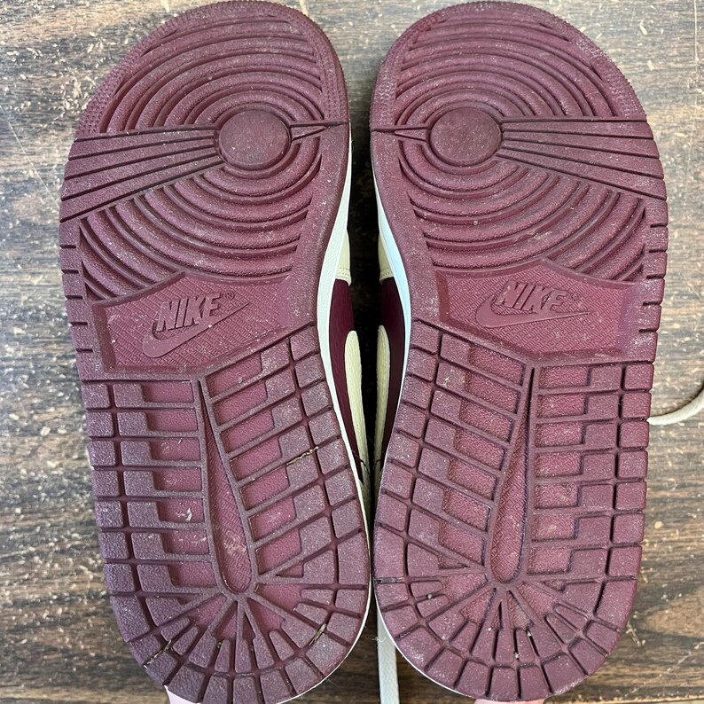 May include: Pair of Nike sneakers, showcasing the soles. The soles are a deep burgundy color with a textured pattern and the Nike logo. The uppers are a cream color with burgundy accents.