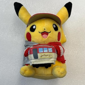 May include: A yellow Pikachu plush toy wearing a red cap, scarf, and a silver jacket. The plush holds a San Francisco cable car. The cable car is red, white, and blue.