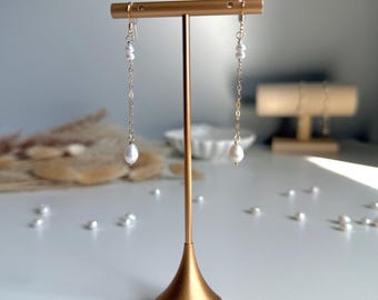 Puede incluir: Expositor de pendientes dorado con dos pares de pendientes colgantes de perlas. Cada pendiente presenta una pequeña cadena con múltiples perlas pequeñas y una perla más grande en forma de lágrima en el extremo. El expositor está sobre una superficie blanca.