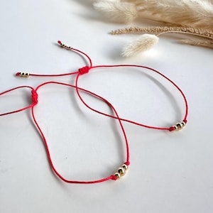 Peut inclure: Deux bracelets en cordon rouge avec des perles dorées. Les bracelets sont liés ensemble et se trouvent sur une surface blanche.