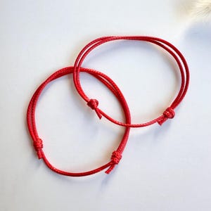 May include: Two red string bracelets tied together in a knot. The bracelets are made of thin, red cord.
