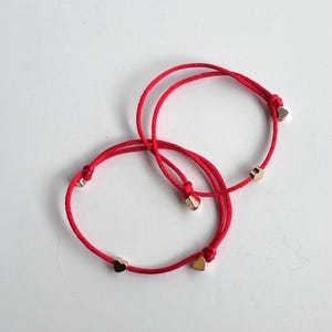 May include: Two red string bracelets with gold heart charms. The bracelets are tied together in a knot.