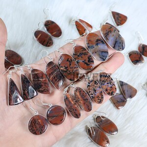Puede incluir: Una colección de pendientes de piedras preciosas con ganchos plateados. Los pendientes presentan varias formas, incluyendo óvalos, lágrimas y triángulos. Las piedras tienen una mezcla de patrones rojos, marrones y negros. Los pendientes se muestran en una mano.