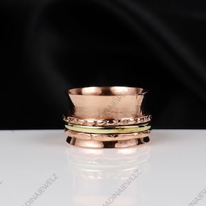 Peut inclure: Une bague rotative de couleur cuivre avec un design texturé. La bague comporte plusieurs bandes, dont une bande dorée et une bande avec un motif d'étoiles. La bague est présentée sur une surface blanche sur fond noir.
