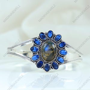 May include: A silver bracelet featuring a central oval-shaped gemstone surrounded by a halo of smaller, deep blue stones. The bracelet's band has a split design with intricate detailing, creating an elegant and eye-catching piece of jewelry.