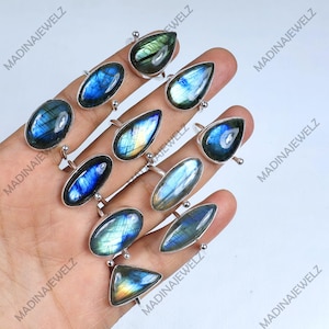 Pode incluir: Uma coleção de anéis de prata com pedras de labradorite iridescentes em várias formas, incluindo ovais, em forma de gota e triangulares. As pedras exibem tons azuis, verdes e amarelos. Os anéis são exibidos em uma mão.
