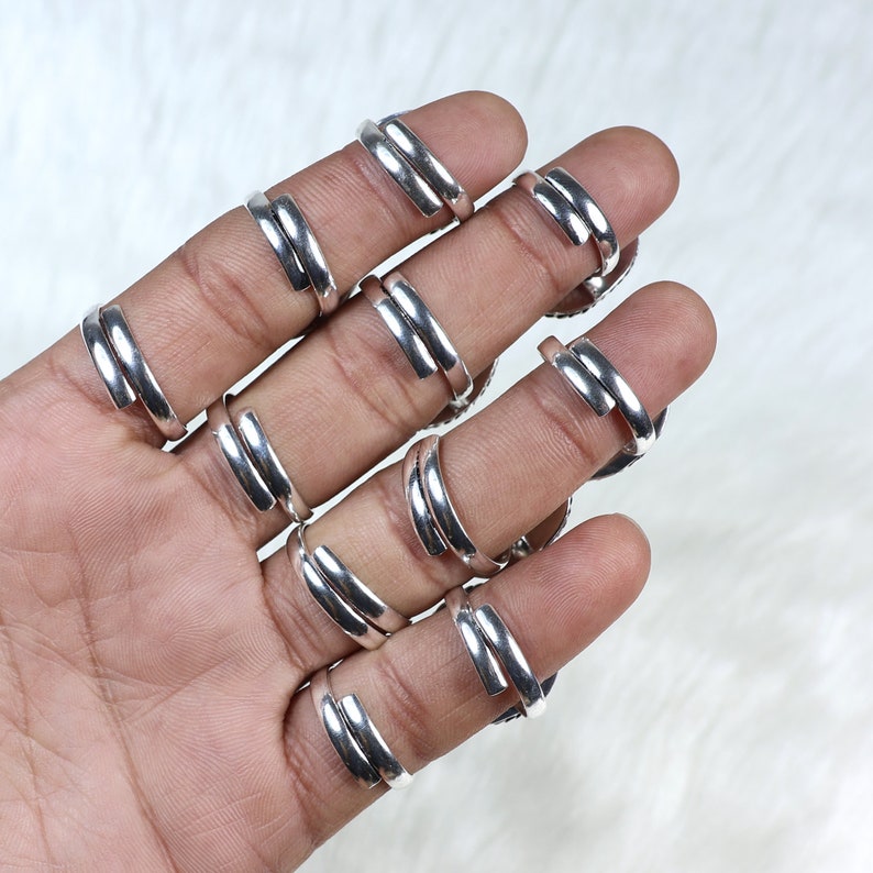 Pode incluir: Uma m&atilde;o exibe v&aacute;rios an&eacute;is ajust&aacute;veis ​​prateados. Cada anel apresenta um design de banda dupla que envolve o dedo. Os an&eacute;is s&atilde;o feitos de um material met&aacute;lico brilhante e s&atilde;o exibidos em um fundo branco macio.