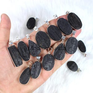 Könnte beinhalten: Eine Sammlung von Anhängern aus schwarzem Turmalin mit silberfarbenen Metalleinfassungen. Die Anhänger haben verschiedene Formen, darunter ovale, rechteckige und tropfenförmige Designs. Die Anhänger werden auf einer weißen Oberfläche präsentiert.